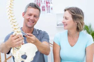 An adult consulting with a health professional and pointing to pain in the shoulder while seated in a calm clinic room.
