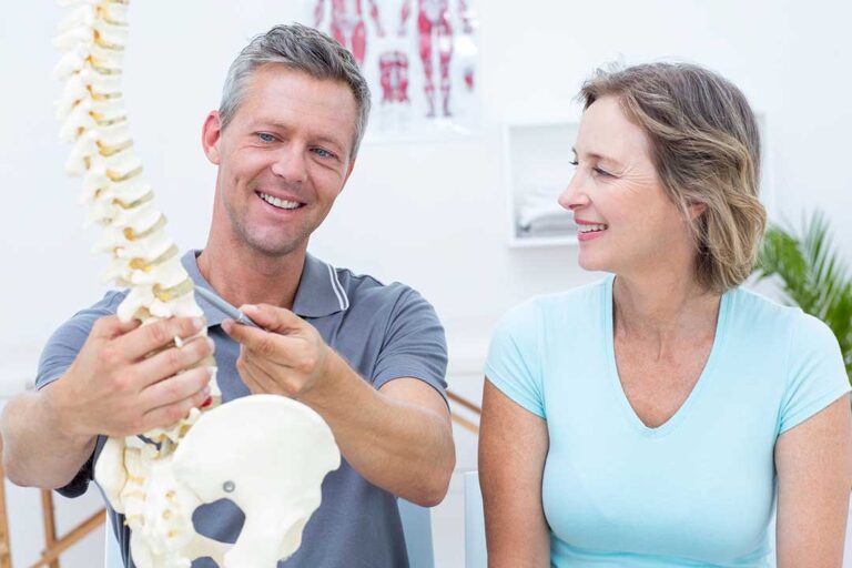 An adult consulting with a health professional and pointing to pain in the shoulder while seated in a calm clinic room.